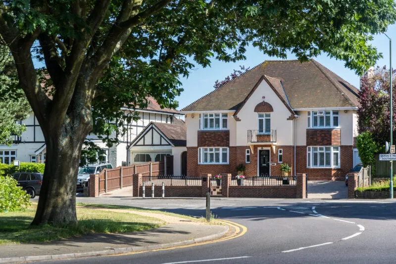 Large detached brick and render house behind a mature tree at a quiet road junction in Queen's Park on a sunny day