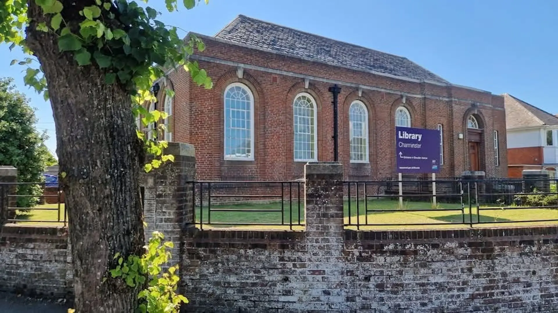 Red-brick Charminster Library with tall arched windows and a purple Library sign behind a low wall