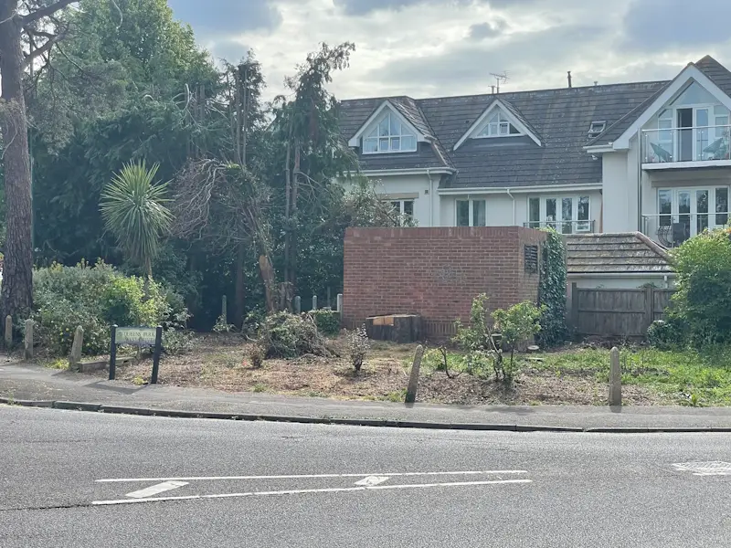 Cleared roadside green space with tree stumps, cut shrubs and a low brick wall in front of a block of flats