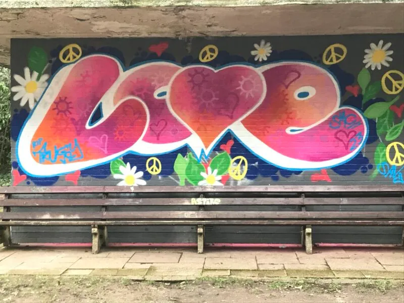 Colourful 'Love' graffiti mural with hearts, daisies and peace symbols behind a wooden bench in a park shelter