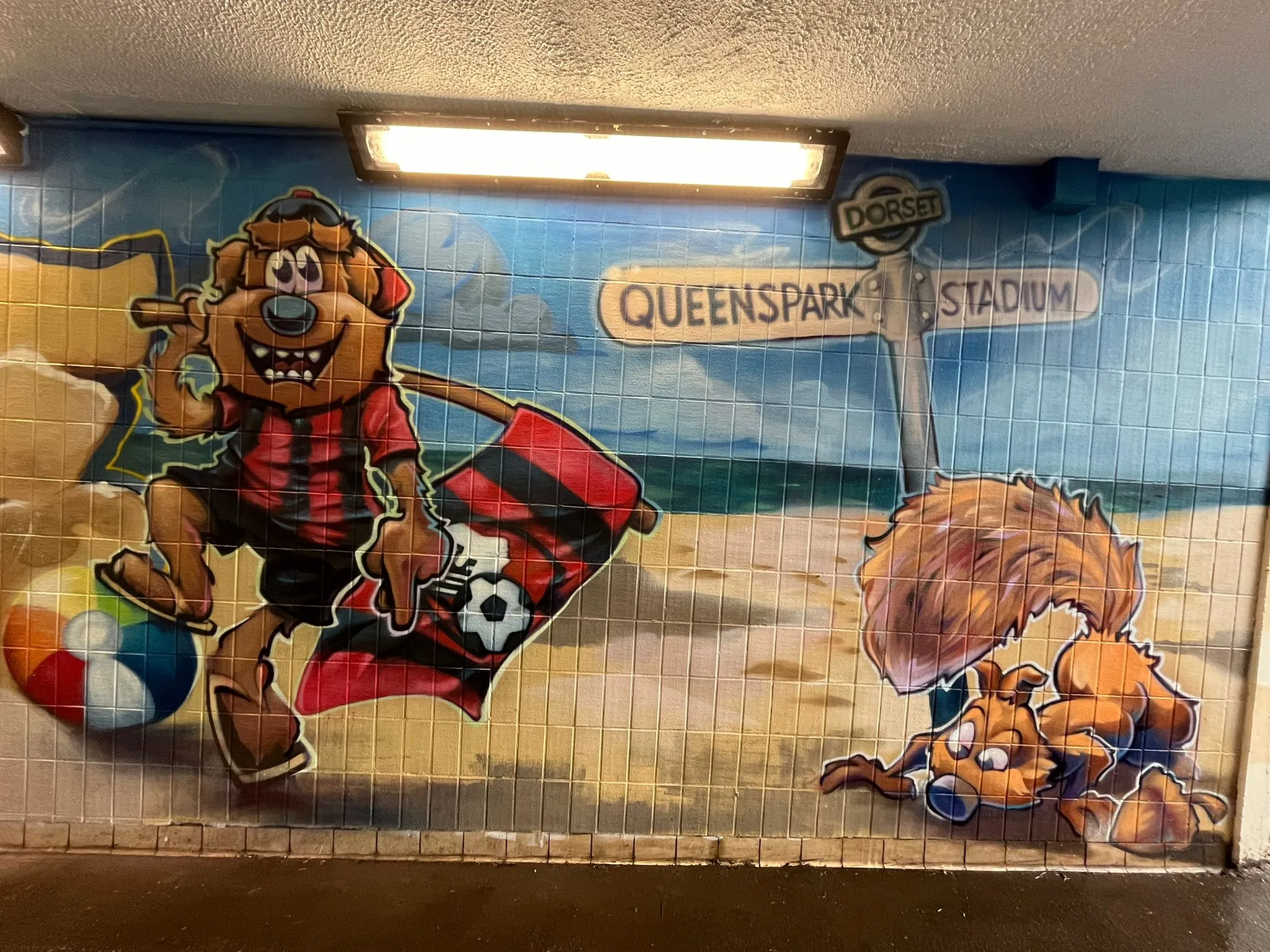 Underpass mural of cartoon animals on a beach beside a signpost reading Queenspark and Stadium