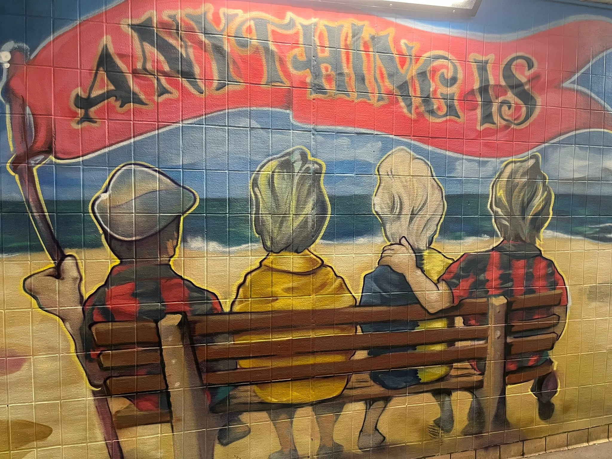 Underpass mural of four people on a beach bench facing the sea, one holding a red flag reading 'Anything is'