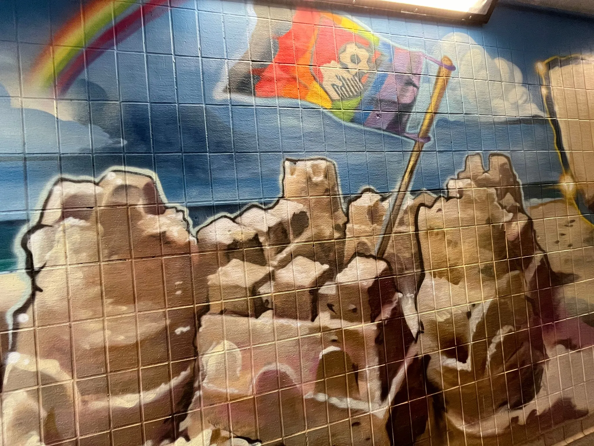 Underpass mural of a sandcastle flying a rainbow flag under a blue sky with a rainbow