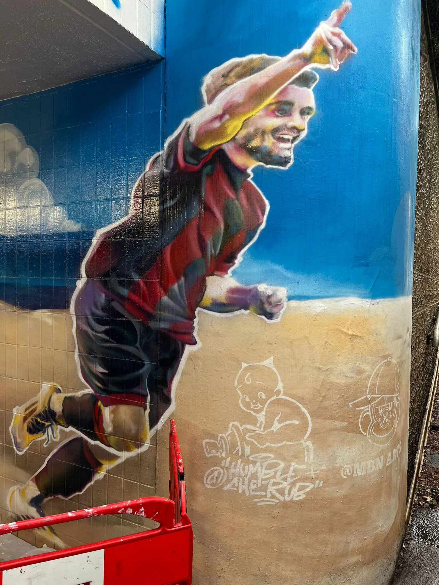 Mural of a footballer in a red and black kit celebrating on a beach, signed Humble Cherub and MBN Arts