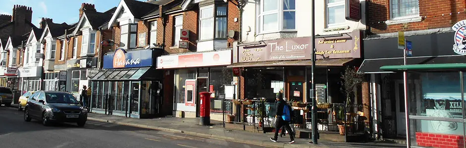 Shopfronts and restaurants along Charminster high street, with a pedestrian passing a red postbox