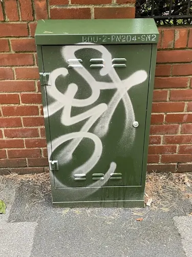 Green street cabinet against a brick wall, defaced with white spray-painted graffiti tags