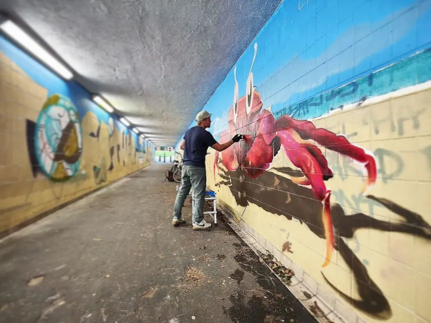 Artist spray-painting a large red crab mural on the tiled wall of a pedestrian underpass