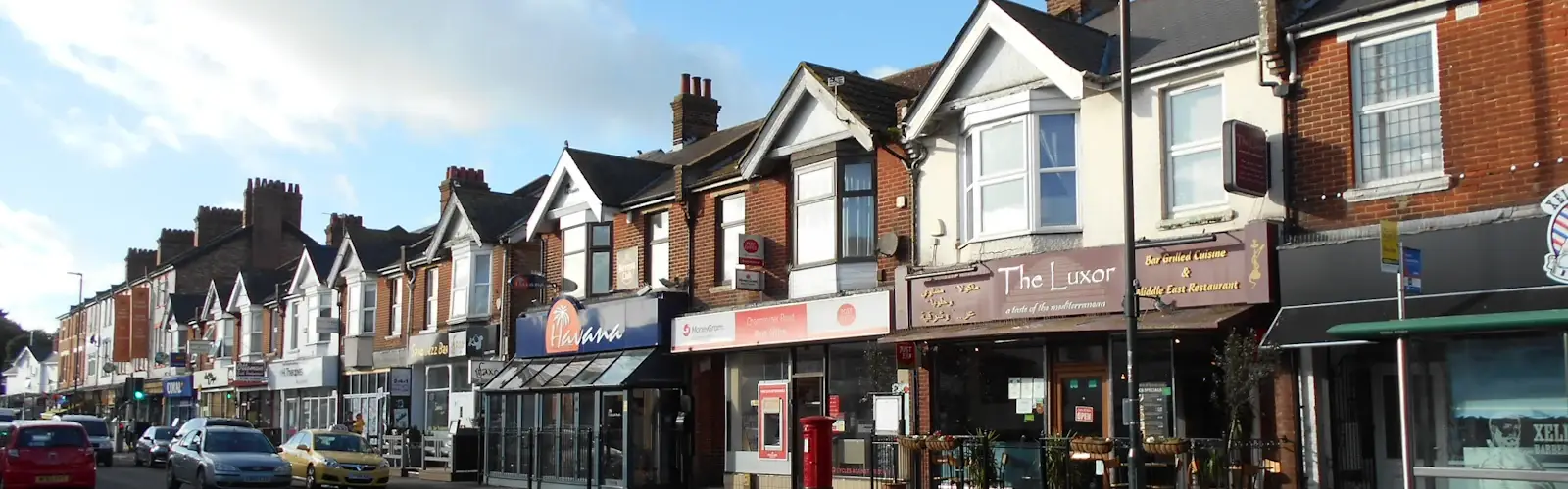 Shops along Charminster Road, including a post office and restaurants, on a sunny day