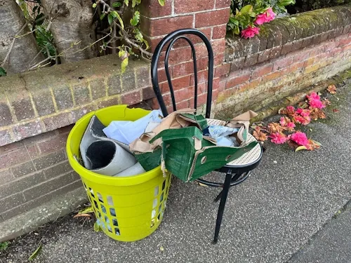 Fly-tipped laundry basket of rubbish and a broken chair left against a garden wall on the pavement