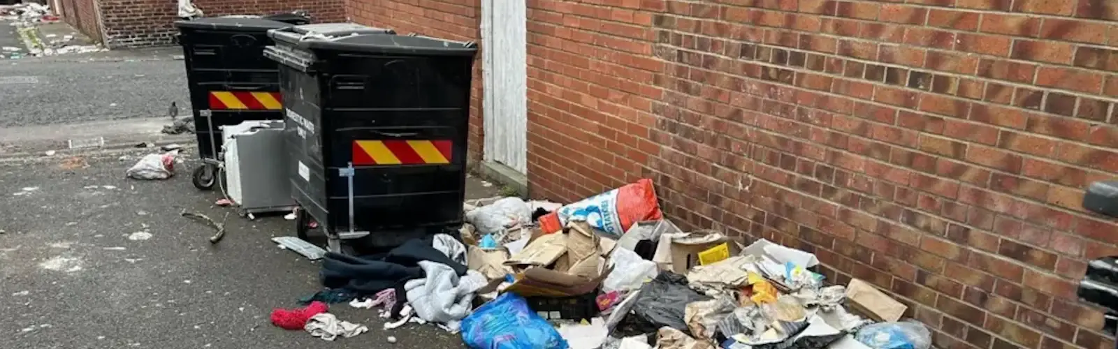 Fly-tipped rubbish, bags and clothing dumped beside communal bins in an alleyway